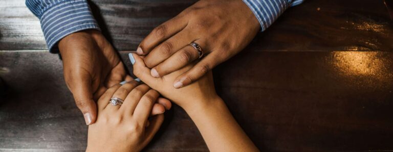 Four hands with wedding rings holding each other's hands protectively.