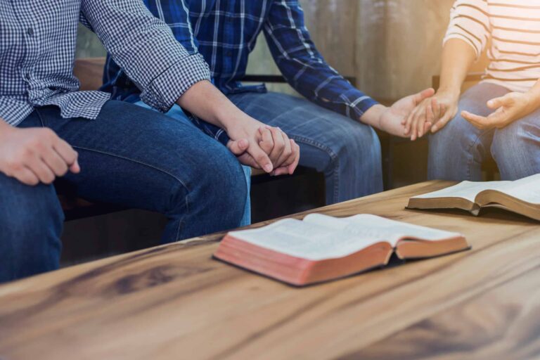 Three people holding hands praying with bible on table in front of them. demonstrating religious practice.