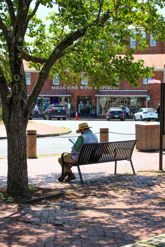 Quiet shady bench with a man wearing a hat is reading an ipad. Mills Fine Wine & Spirits store and sign behind. Anapolis, MD