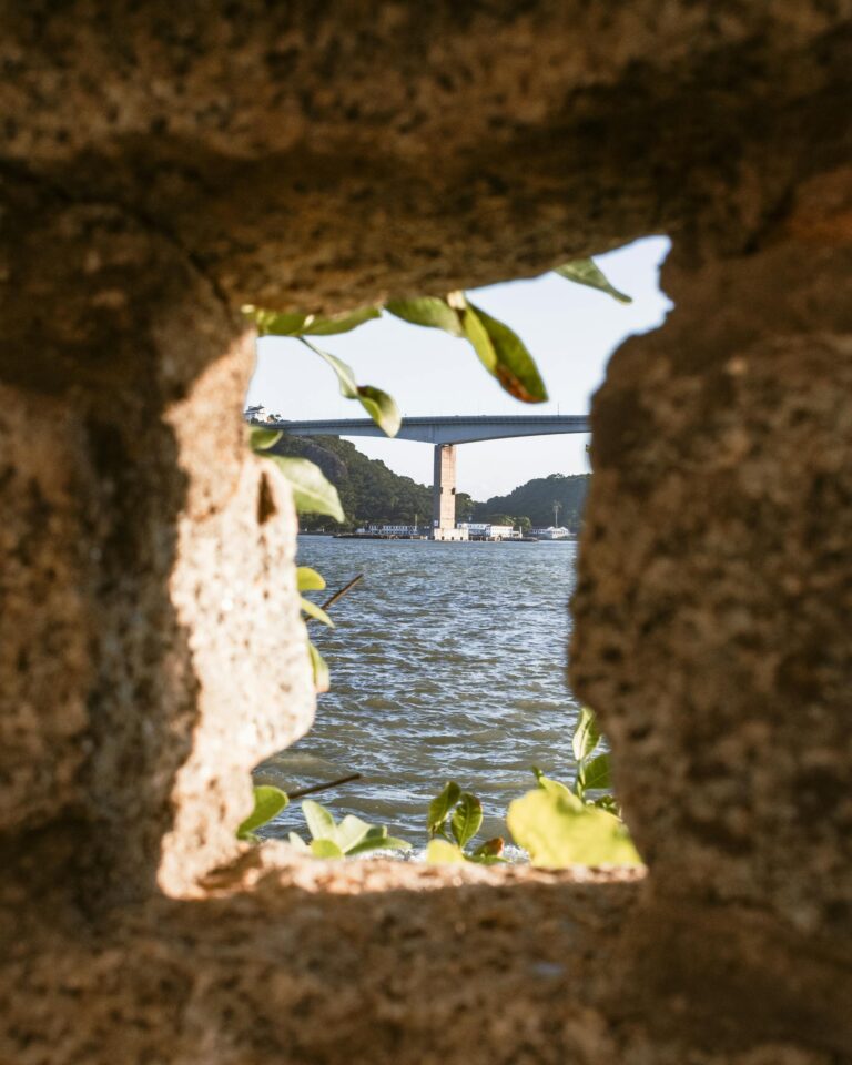A beautiful view of a bridge over water framed through a stone window with leafy greens.