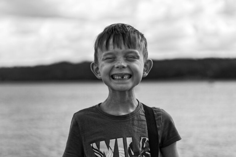 A cheerful young boy smiles broadly by a lake, captured in a black and white portrait.