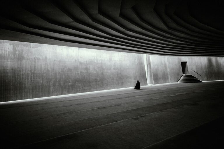 A lone person meditates in a vast, modern, minimalist indoor hall with dramatic architecture.