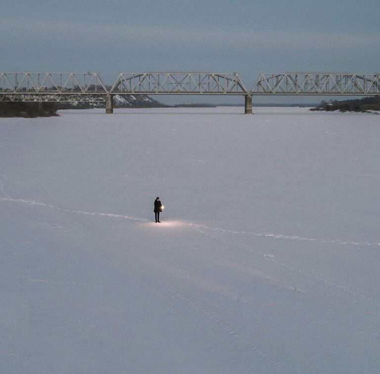 A lone person walks across a snow-covered frozen river, carrying a lantern, beneath a distant bridge.
