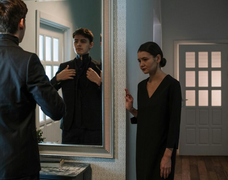 A man adjusts his suit in front of a mirror with a woman looking on in an elegant room.