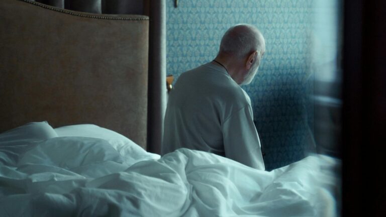 A man sits pensively by the window in a dimly lit bedroom, creating a mood of solitude and reflection.