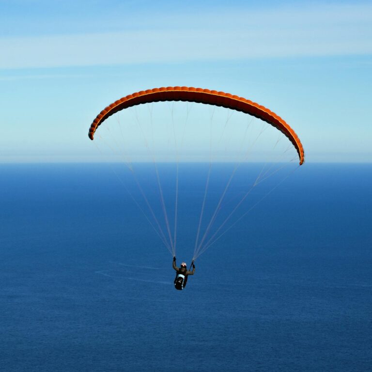 A person paragliding with an orange chute over a vast ocean under clear blue skies.