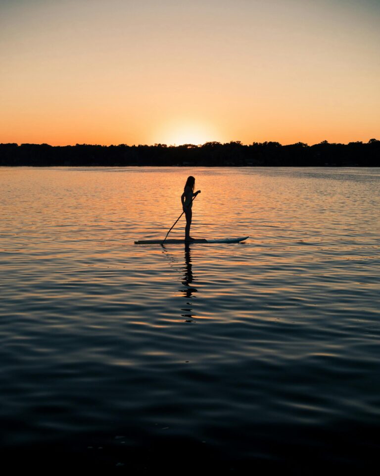 A silhouette of a woman paddleboarding at sunset on a tranquil Minnesota lake, backlit by the setting sun.