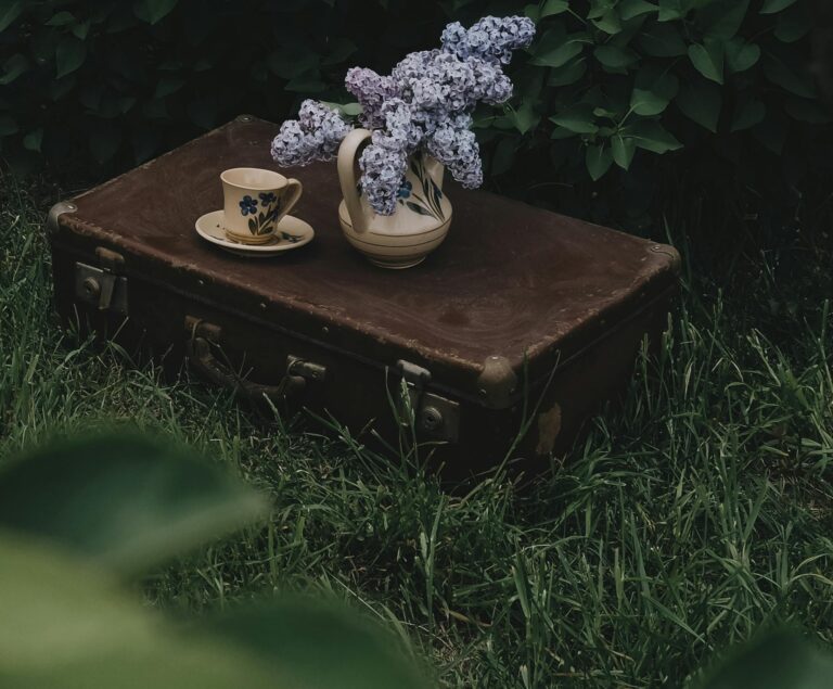 A vintage suitcase topped with a floral vase and tea cup outdoors, embracing rustic charm.
