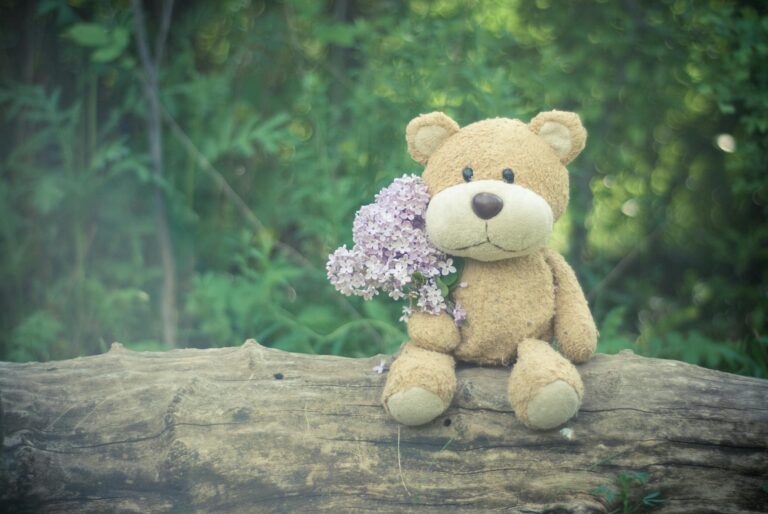 Adorable teddy bear with lilac flowers on a log in a serene forest setting.