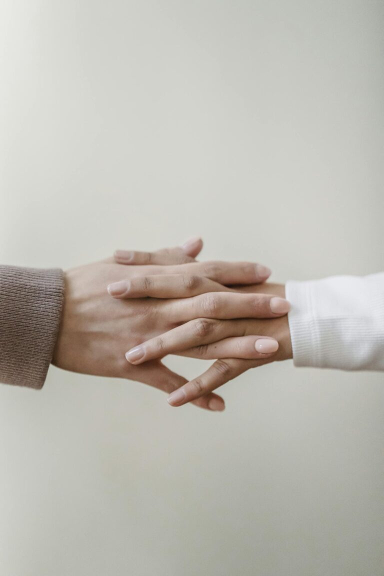 Crop unrecognizable female friends in casual outfit holding hands in light apartment near white wall