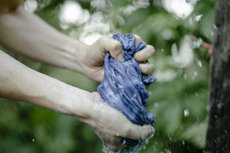 Hands of faceless person squeezing wet blue fabric under water drops while standing on street near green plants on blurred background