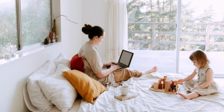Side view of cute toddler girl sitting on bed barefoot and playing with colorful wooden blocks while mother using laptop in bed enjoying morning coffee