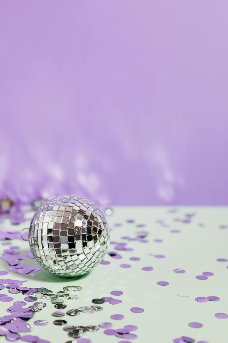 Silver disco ball amid vibrant purple and green confetti with a light purple background