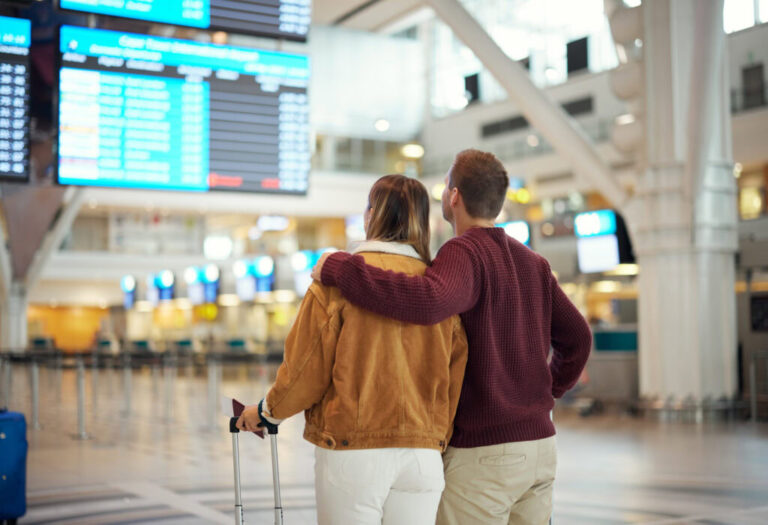 A couple in an airport looking up at the flight arrivals and departures. They carry luggage but only the handles are showing. He is wearing a maroon sweater and she has on a tan jacket with white color. His arm is around her.