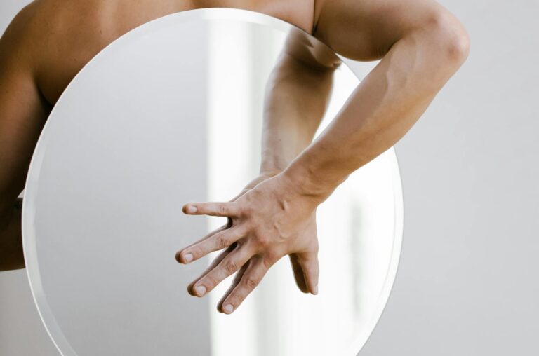 A bald man from behind holding a round mirror against a white wall indoors.